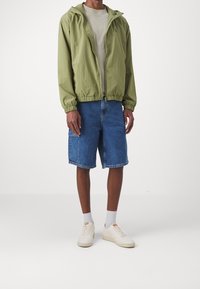 Man som bär en ljusgrön zip-up jacka, beige t-shirt, blå denim cargo shorts, vita strumpor och vita sneakers, stående på ett vitt golv.