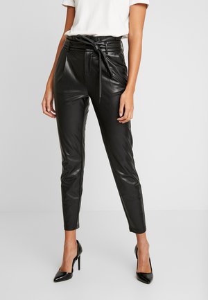 Vrouw draagt zwarte leren broek met hoge taille en een ceintuur, gecombineerd met zwarte hoge hakken en een wit T-shirt met korte mouwen.