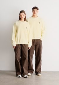 Šviesiai geltoni džemperiai su subtiliu logotipu, derinami su laisvomis rudos spalvos kelnėmis, papildyti pilkais ir baltais sportbačiais. Paprastas, modernus dizainas.