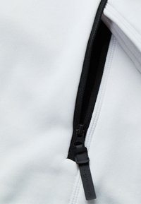 Valkoinen kangas, jossa on musta vetoketju ja teksturoitu pinta. Vetoketjun nyppylässä on brändäys. Muotoilu on minimalistinen ja toiminnallinen.