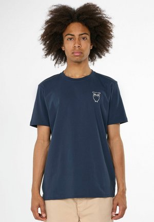 Jeune homme aux cheveux bouclés portant un t-shirt bleu marine avec un petit motif de chouette blanche sur la poitrine, debout devant un fond blanc uni.