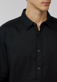 Chemise noire à boutonnières avec un tissu texturé, col traditionnel et patte de boutonnage sur le devant avec des boutons foncés.