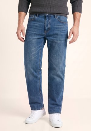 Homme portant un jean bleu moyen, des baskets blanches et un pull tricoté gris foncé à manches longues, debout, les bras détendus.