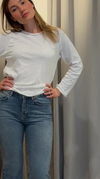 Ung kvinne med langt hår iført en hvit langermet skjorte og blå jeans, stående med hendene på hoftene foran et nøytralt gardinbakgrunn.