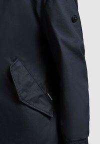 Rękaw ciemnoniebieskiej kurtki z ukośną klapą kieszeni i widocznym czarnym guzikiem na górnej części ramienia na jasnym tle.