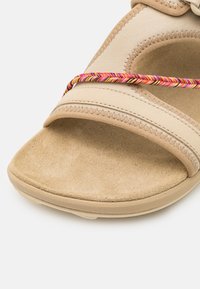 Beige sandaler med en myk semsket såle og elastiske stropper med en flerfarget flettet detalj over den øvre delen.