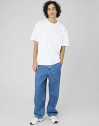 T-shirt en coton blanc avec une poche sur la poitrine, associé à un jean en denim baggy bleu clair. Les chaussures sont des baskets blanches avec des accents gris.