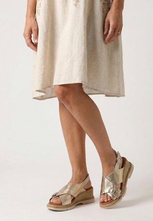 Beine mit beige-metallischen Keilsandalen mit Schnallen und einem hellbeigen kniehohen Kleid, stehend vor einem einfarbigen Hintergrund.