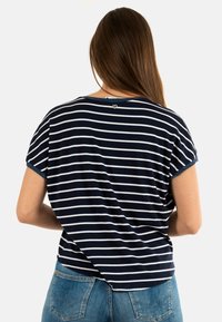 Camiseta a rayas en azul marino y blanco, con mangas cortas, cuello redondo y un pequeño detalle en forma de corazón en la parte trasera. Combinada con vaqueros azules.