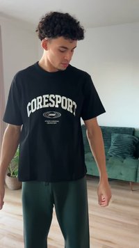 Fekete póló fehér, ívelt "CORESPORT" felirattal és további márkajelzésekkel; sötétzöld, bő szabású nadrággal párosítva.