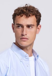 Jonge man met krullend bruin haar, die een lichtblauw overhemd met knopen over een wit T-shirt draagt, terwijl hij naar links kijkt tegen een effen achtergrond.