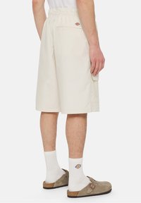 Mies, joka käyttää vaaleanbeigejä cargo-shortsia, valkoisia sukkia logolla ja beigen nubukista valmistettuja avokärkisiä sandaaleja, seisoo yksinkertaista taustaa vasten.