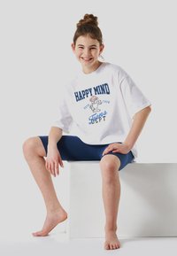 White oversized T-shirt with blue text and cartoon graphic. Worn with navy leggings, seated on a white cube. Barefoot, smiling.
