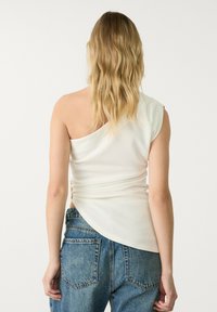 Haut crème à une épaule en tissu lisse. Présente un design drapé avec un détail froncé sur le côté et un ourlet légèrement asymétrique.
