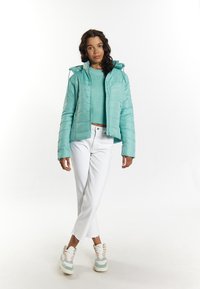 Jachetă puffer teal cu glugă, închidere cu fermoar și textură matlasată, asortată cu un pulover verde deschis scurt și blugi albi. Încălțăminte cu detalii accentuate.