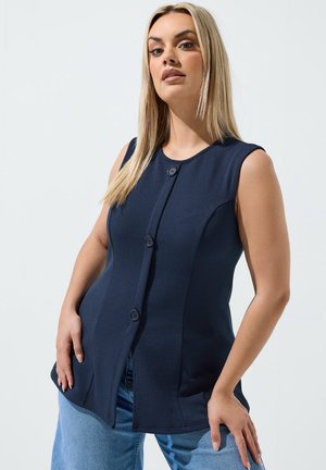 Kvinde med glat blondt hår iført en ærmeløs marineblå knapbluse og lyseblå jeans, poserer med hænderne hvilende på hofterne.