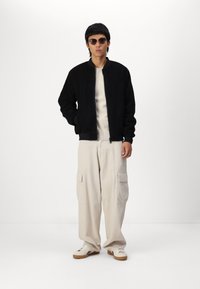 Jachetă bomber din fleece neagră cu fermoar în față, pantaloni cargo bej și adidași albi cu tălpi maro. Accesorizată cu ochelari de soare rotunzi.