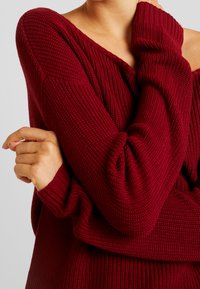 Maglione lavorato a maglia bordeaux con vestibilità ampia, scollatura larga, righe testurizzate e maniche lunghe. Presenta cuciture delle spalle scese e polsini ripiegati.