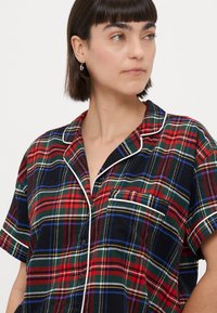 Red and green plaid short-sleeve shirt with white piping, featuring a chest pocket and a relaxed fit, made from a soft fabric.