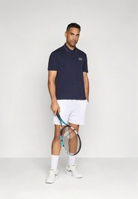 Polo azul-marinho com gola e logótipo, combinado com calções brancos. O modelo segura uma raquete de ténis e usa sapatos e meias desportivas brancas.