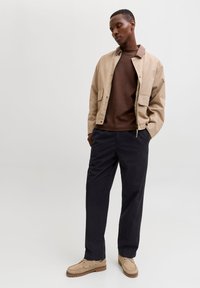 Μπεζ μπουφάν με καφέ γιακά, καφέ φούτερ από μέσα, και σκούρο παντελόνι. Ταμπά παπούτσια με σχέδιο μοκασίνι. Ουδέτερο φόντο.