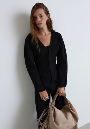 Frau mit welligem Haar, die ein schwarzes Langarmshirt und Hose trägt und eine große beige Wildledertasche mit Fransen vor einer weißen Wand hält.