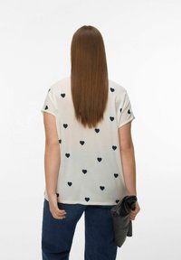 Vrouw met lang, stijl haar, gekleed in een wit shirt met zwarte harten, blauwe spijkerbroek, houdend een opgevouwen zwarte leren clutch achter haar rug.