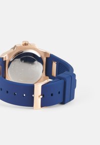 Correa de reloj de silicona azul con un cierre de acero inoxidable en color oro rosa, que presenta una caja redonda y un fondo transparente.