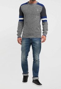 Pull gris en maille avec rayures bleues et blanches sur les manches, col rond, associé à un jean bleu et des baskets noires.