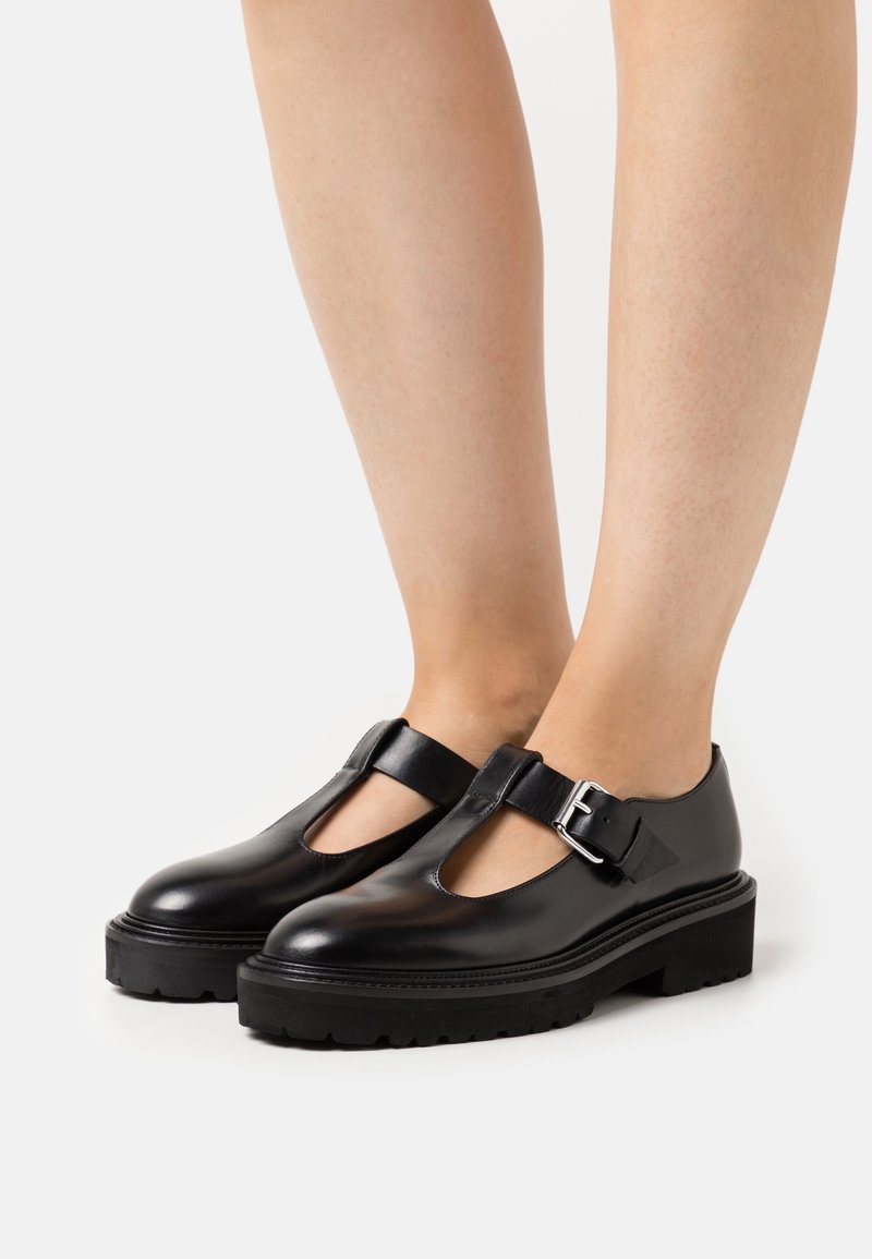 Chaussures Mary Jane en cuir noir avec semelles crantées épaisses et lanières à boucle argentée portées pieds nus sur fond blanc.