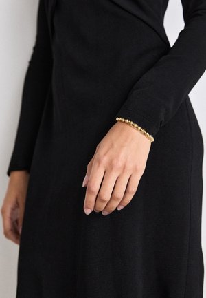 Main portant un bracelet en perles dorées reposant contre le tissu noir d'une robe à manches longues.