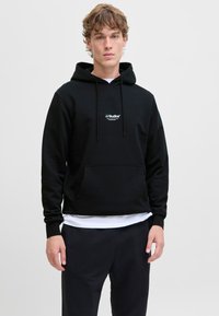 Zwarte hoodie van zacht materiaal met een voorzak, trekkoordkap en wit "JJ Studios" logo op de borst. Draagt over een wit shirt.