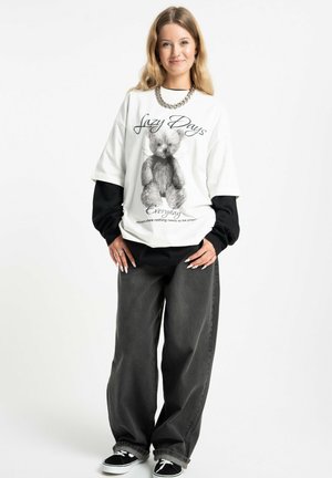 Młoda kobieta ubrana w warstwowe czarno-białe ubrania codzienne z t-shirtem z grafiką misia pluszowego i czarnymi, szerokimi jeansami, uśmiechająca się do kamery.