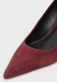 Gros plan sur une chaussure femme en daim bordeaux à bout pointu, intérieur noir et détail de bord festonné.