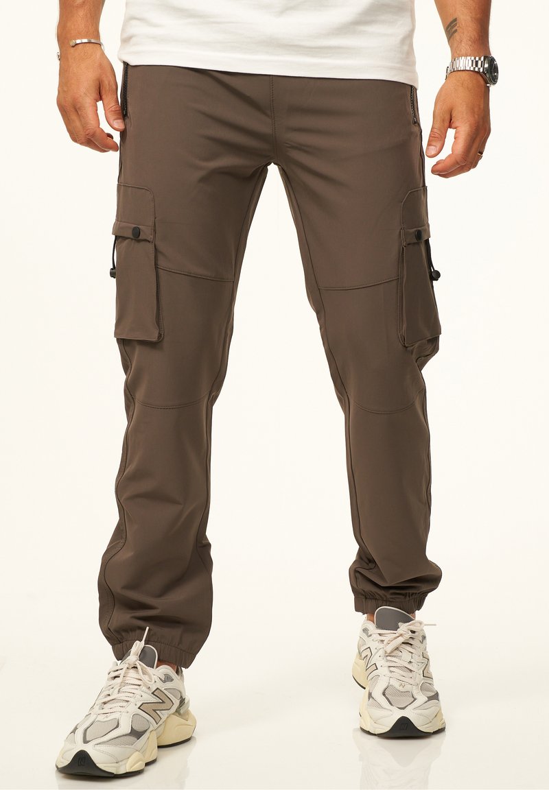 Braune Cargo-Hosen mit mehreren Seitentaschen, elastischen Bündchen und einer glatten Textur. Getragen mit hellen, klobigen Sneakern.