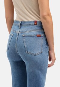 Lichtblauwe spijkerbroek met hoge taille, achterzakken en een rood label op de tailleband. De stof lijkt zacht met een gladde textuur.