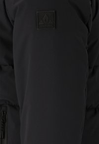 Svart vadderad jackärm med svart Whistler-märke och synlig dragkedjeficka på nedre sidan.