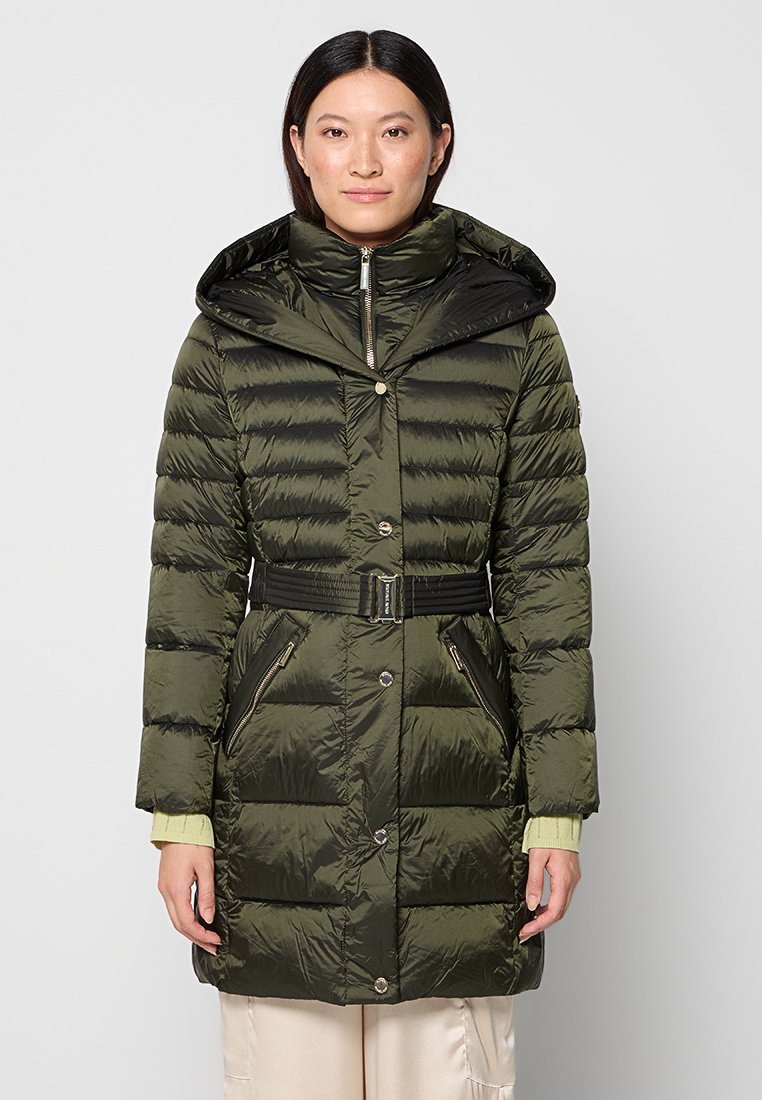 Frau in einem dunkelgrünen, mit einem Gürtel versehenen Daunenmantel mit hohem Kragen und Reißverschlusstaschen, die vor einem schlichten hellen Hintergrund steht.