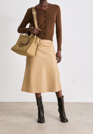 Personne portant un cardigan en maille torsadée marron, une jupe midi beige, des bottines noires à talons, tenant un sac cabas Coach beige avec bandoulière.
