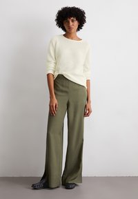 Hellgelber strukturierter Pullover mit weitem Ausschnitt, kombiniert mit olivgrünen, weit geschnittenen Hosen mit seitlichen Schlitzen und schwarzen Slippers.