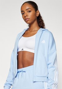 Svetlo modra zip-up kapuca z belimi črtami na rokavih, nosi se preko bele športne nedrčke. Material se zdi mehak; ima sproščen in neformalen dizajn.