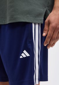 Κοντινό ενός ατόμου που φοράει σορτς Adidas σε ναυτικό μπλε με λευκές ρίγες και λογότυπο, σε συνδυασμό με σκοτεινό πράσινο πουκάμισο, με ορατό το δεξί χέρι.