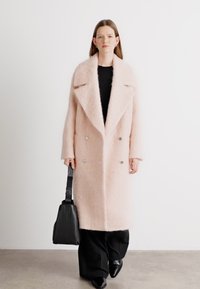 Casaco longo rosa e fofinho com um colarinho largo, botões duplos e bolsos frontais; usado sobre uma blusa preta e combinado com calças pretas e uma bolsa.