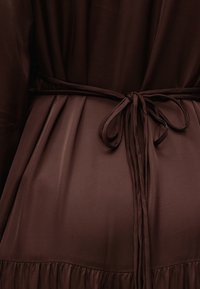Robe en tissu marron foncé nouée à la taille avec une fine ceinture formant un simple nœud, montrant de doux plis et plis religieuses sur la partie inférieure.