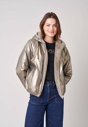 Jonge vrouw die lacht, draagt een metalen capuchonjas met bontvoering, een zwart shirt met ontwerp en een blauwe spijkerbroek met hoge taille.