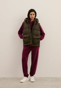 Brązowa kamizelka puchowa z kapturem, z dwoma przednimi kieszeniami. Nosi się ją na bordowy hoodie i dresowe spodnie. Białe sneakersy dopełniają stylizację.