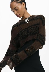 Barna, rövidített kötött pulóver, texturált csíkokkal, nyitott kötésű részletekkel és hosszú ujjal. Fekete testhezálló ruhadarabra viselve.