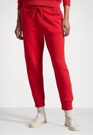 Personne portant un pantalon de jogging rouge avec cordon et logo bleu, des chaussettes blanches et des baskets blanches, debout sur un fond clair uni.