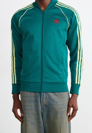 Mężczyzna w turkusowej kurtce Adidas na zamek z żółtymi paskami na rękawach i czerwonym logo, w połączeniu z wyblakłymi niebieskimi dżinsami.