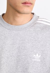 Sivi dukserica s okruglim izrezom, napravljena od pamuka, s bijelim Adidas logotipom i tri bijele pruge na ramenima, glatke teksture.